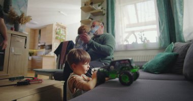 Baba iki çocukla oturuyor, biri yeşil bir oyuncak kamyonla oynuyor, diğeri de sıcak bir aile salonunda, modern ve sıcak bir dekorla izliyor.