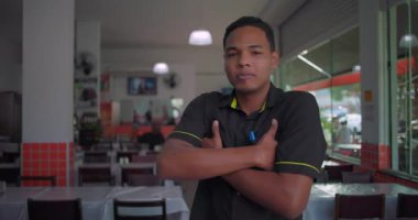Yemek alanında kolları çapraz duran Latin restoran çalışanı, masalar ve pencere ışıklarıyla dolu parlak bir şekilde poz verirken kendine güvenen bir ifade.