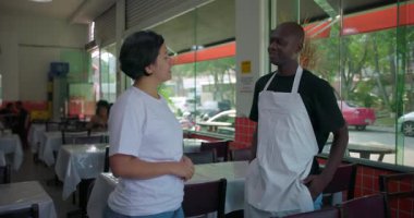 Restoranda konuşurken kadın yöneticiyi dinleyen Afro-Amerikan garson, doğal ışıkla günlük yemek alanının penceresinin yanında duruyor.