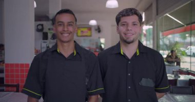 İki gülen restoran çalışanı yemek alanında duruyor, biri önlüklü, kapalı mekan sahnesi masaları, sandalyeleri ve arka planda büyük pencereleri olan.