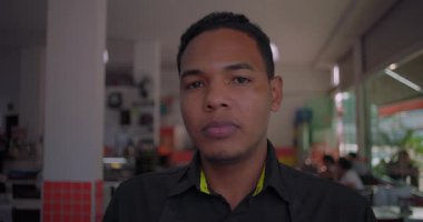 Nötr ifadeli Latin lokantası çalışanı, yemek alanında cam pencereler ve yumuşak iç aydınlatmayla duruyor.