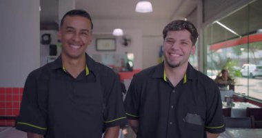 Yan yana duran iki gülümseyen restoran çalışanı kameraya bakıyor. Kapalı yemek odası, fayanslı duvarlar ve arka planda büyük pencereler var.