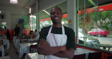 Restoranda duran Afro-Amerikalı adam, kollarını kavuşturmuş ve güvenle gülümsüyor, cam pencereli ve arka planda oturan müşterileri olan kapalı bir yemek salonunda.