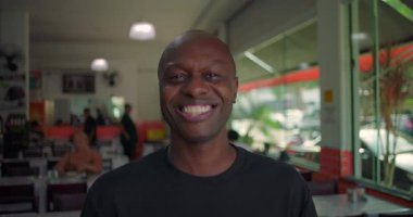 Gülümseyen Afro-Amerikalı bir adam sıradan yemek alanında kameraya bakıyor. Oturan insanlarla neşeli bir ruh hali ve büyük restoran pencerelerinin önünde sokak manzarası var.