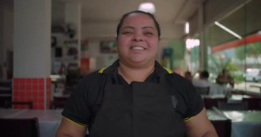 Yemek alanında kolları çapraz duran Latin restoran çalışanı, güvenli bir şekilde gülümsüyor. Parlak, modern bir restoranda doğal pencere ışığında.