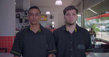 İki restoran çalışanı yemek alanında yan yana duruyor, biri önlük giyiyor, ikisi de ciddi görünüyor, arka planda masalar ve cam duvar görünüyor.