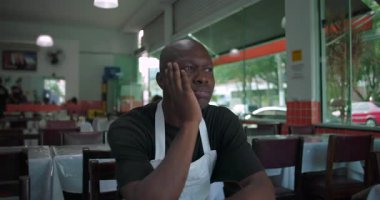 Afro-Amerikan bir adam restoranda el ele dinleniyor, düşünceli ve biraz endişeli bir ifadeyle kapalı yemek alanında yakın çekim