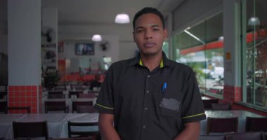 Lokanta yemek alanında duran Latin garson, tarafsız bir ifade, elleri yanda, cebinde kalemle üniforma giyiyor, masalarla çevrili.