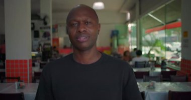 Afro-Amerikalı bir adam sakin bir şekilde günlük restoranın içinde kameraya bakıyor. Tarafsız bir ifadeyle yakın plan portresi, arka planda yemek yiyen insanlar.