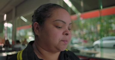 Latin kadın restoran çalışanı pencerenin yanında dururken sessizce aşağı bakıyor, gündelik yemek mekanında kısa bir mola sırasında düşüncelere dalmış.