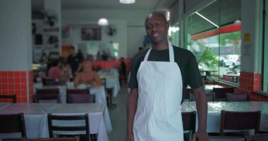 Lokantada gülümseyen beyaz önlük giyen Afro-Amerikalı bir adam. Elleri yan yatmış masaların yanında duruyor.