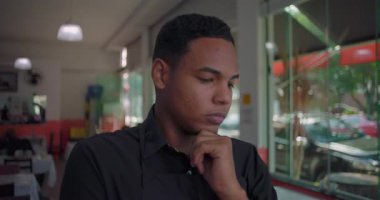 Elleri çenesinde aşağı bakan Latin restoran çalışanı modern yemek alanında doğal pencere ışığıyla derin düşüncelere dalmış.