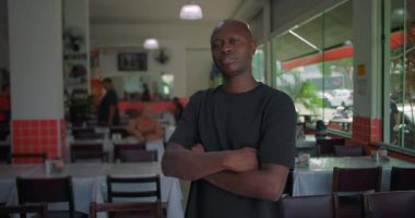 Sıradan bir restoranda duran Afro-Amerikalı adam kollarını kavuşturmuş, kendine güvenen ve ciddi bir ifade takınıyor, cam pencereler ve arka planda oturan lokantalar.