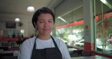 Lokanta yemek alanında kolları çapraz duran Latin garson, kameraya bakarken yumuşak bir ifade, masalar ve büyük cam pencerelerle çevrili.