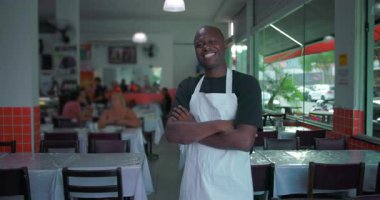 Beyaz önlüklü gülümseyen Afro-Amerikalı adam rahat yemek ortamında rahat ve samimi bir şekilde kollarını kavuşturdu.