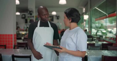 Afro-Amerikalı çalışan ve bayan yönetici tablet üzerinde konuşuyorlar, her ikisi de ekranda parlak restoran ortamında dikilirken
