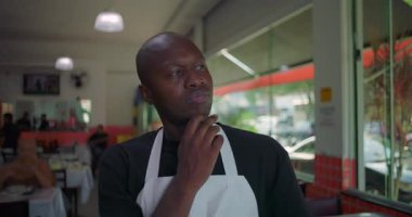 Restorandaki Afro-Amerikalı adam, eli çenesinde dalgın bir şekilde yemek masasının önünde duruyor, müşterileri ve personeli hafif bir odak noktasında.