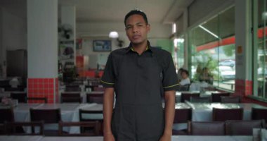 Yemek alanında kolları çapraz duran Latin restoran çalışanı, güneşli restoran ortamında rahat duruş ve nötr ifade