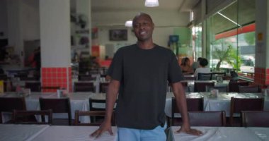 Rahat duruşu olan, sandalyelerde dinlenen, kendinden emin bir ifadeyle kameraya bakan, arka plandaki masalara bakan, sıradan bir restoranda duran Afrikalı Amerikalı adam.