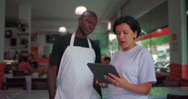 Afro-Amerikan restoran çalışanı kadın yöneticiyle tableti inceliyor, iş yerinde kapalı alanda yapılan açık oturum sırasında yan yana duruyor.