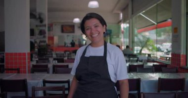 Lokanta yemek alanında sıcak bir şekilde gülümseyen Latin garson, elleri sımsıkı bağlanmış ve kolları farklı anlarda kavuşturulmuş, masalar ve doğal pencere ışığı ile çevrili.