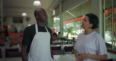 Afro-Amerikan restoran çalışanları, günlük yemek alanında kadın yöneticiyle konuşuyor. İkisi de pencere kenarındaki rahat etkileşim sırasında gülümsüyor.