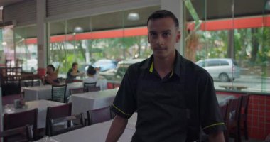 Önlüklü restoran garsonu yemek alanında tek başına duruyor, ciddi görünüyor, boş masalar ve arka planda pencere yansımaları var.
