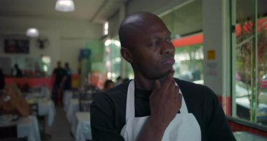 Afro-Amerikalı bir adam, düşünceli bir ifadeyle restoranda dikiliyor, yüzünü yana çevirirken çenesine dokunuyor, doğal ışık ve arka planda oturan lokantalar.