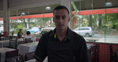 Önlüklü erkek restoran çalışanının doğrudan kameraya, ciddi ifadeye, cam pencerelere ve restoranın dışındaki trafiğe bakması.