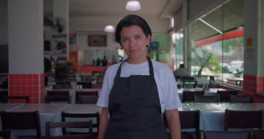 Latin garson restoranın yemek salonunda duruyor, sakin ve tarafsız bir ifadeyle doğrudan kameraya bakıyor, masalarla ve doğal ışıklarla iç mekan ayarları yapıyor.