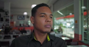 Latin lokantası çalışanı çerçeveden dışarı bakıyor. Büyük pencereli, parlak yemek alanında sakin ve yansıtıcı bir ifade.