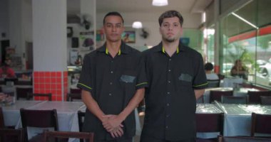 İki erkek restoran çalışanı yemek alanında yan yana duruyorlar, elleri önde katlanmış, ciddi görünüyorlar, kapalı mekan sahneleri plastik ambalajlarla kaplı.