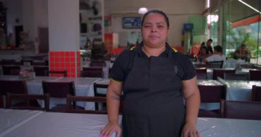 Latin lokantası çalışanı yemek alanında nötr bir ifade takınıyor, modern kapalı mekan ortamında parlak doğal ışık altında önlük giyiyor.