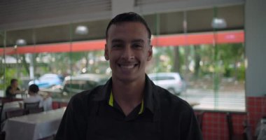 Önlüklü restoran çalışanı yemek alanındaki kameraya gülümsüyor, plastik kaplı masalar ve arka planda görünen cam pencereler.