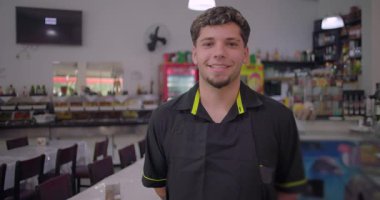 Gülümseyen restoran garsonu sıradan bir restoranın içinde kameraya poz veriyor masaların ve büfe alanının önünde duruyor ve arka planda buzdolapları var.