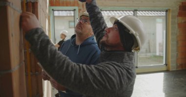 İnşaat işçilerinin yakın çekimde tuğla duvar ve meslektaşlarının gözlemlemesi, inşaat alanında büyük pencereli takım çalışması devam ediyor