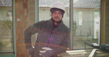 Kendine güvenen inşaat işçisi, kısmen inşa edilmiş bir yapının çalışma tezgahının yanında elleri ve kalçaları üzerinde poz veriyor, güvenlik donanımı, takım çalışması ve inşaat alanında gurur takıyor.