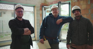 Tamamlanmamış binada inşaat ekibi, proje planlarını ellerindeki planlar, takım çalışması, güvenlik kaskları, işbirliği ve inşaat ayrıntılarına odaklanıyor.