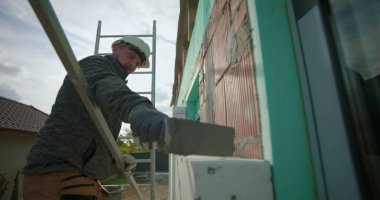 Dış duvardaki yalıtım panelini malayla temizliyorum, inşaat işçisi, tuğla inşaatında çalışıyorum, miğfer ve eldivenle merdiven üzerinde duvar işçiliği yapıyorum.