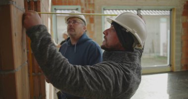 İnşaat işçilerinin yakın çekimde tuğla duvar ve meslektaşlarının gözlemlemesi, inşaat alanında büyük pencereli takım çalışması devam ediyor