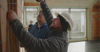 İnşaat işçilerinin yakın çekimde tuğla duvar ve meslektaşlarının gözlemlemesi, inşaat alanında büyük pencereli takım çalışması devam ediyor