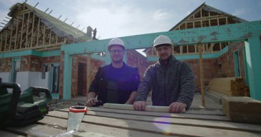 İnşaat alanında duran inşaatçılar, çerçeve altındaki ev, takım çalışması ve işbirliği, ahşap şapkalı iki adam, aletler ve malzemelerle çalışabilir, tamamlanmamış bir ev.