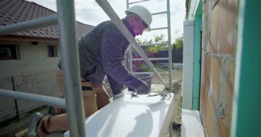 Yalıtım tahtasına yapıştırıcı süren inşaat işçisi, duvar kurulumu, güvenlik kaskı, arka planda merdiven ve yapı iskelesi olan dış yapı projesi için hazırlık yapıyor.