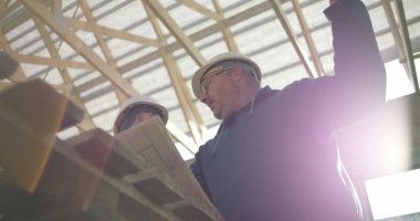 İnşaat danışmanı ve iş arkadaşı proje detaylarını, takım çalışmasını ve rehberliği tartışıyorlar. Her ikisi de arka planda yapısal çerçeveye sahip güvenlik miğferlerinde.