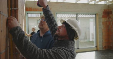 İnşaat işçilerinin yakın çekimde tuğla duvar ve meslektaşlarının gözlemlemesi, inşaat alanında büyük pencereli takım çalışması devam ediyor