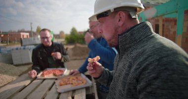 İnşaat alanında pizzayla öğle yemeği arası veren üç müteahhit, gelişigüzel toplanma, takım çalışması ve bağlanma, inşaat halindeki konut projesi.