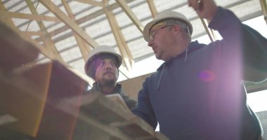 İnşaat danışmanı ve iş arkadaşı proje detaylarını, takım çalışmasını ve rehberliği tartışıyorlar. Her ikisi de arka planda yapısal çerçeveye sahip güvenlik miğferlerinde.