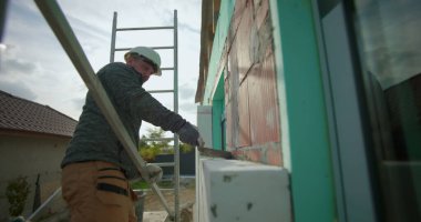 Dış duvardaki yalıtım panelini malayla temizliyorum, inşaat işçisi, tuğla inşaatında çalışıyorum, miğfer ve eldivenle merdiven üzerinde duvar işçiliği yapıyorum.