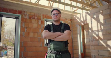 Kendine güveni tam bir inşaat işçisi, kolları çapraz, sıkı bir şapka ve tulum giyiyor, bitmemiş bir binanın içinde duruyor, kararlılık ve profesyonellik sergiliyor.