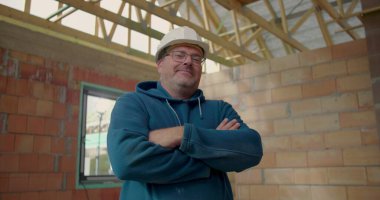 Kollarını kavuşturmuş, kasketli, kısmen inşa edilmiş bir binada duran kendine güvenen bir inşaat işçisi. İşinde profesyonelliği ve gururu somutlaştırıyor.